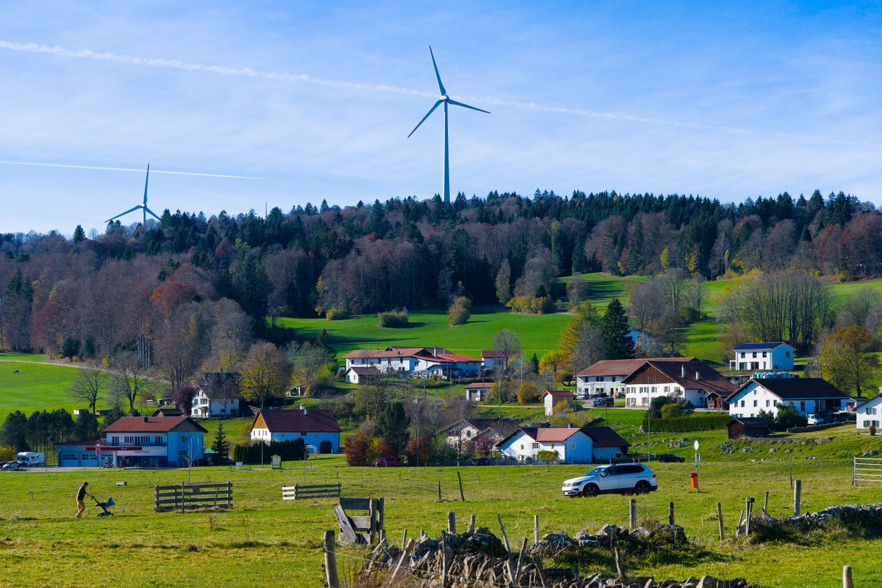 contact-img Picturesque Les Breuleux village with modern wind turbines and lush green fields under a clear blue sky.