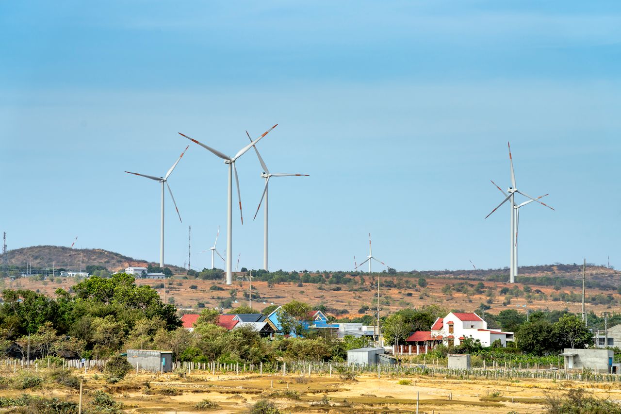 Scenic view of wind generators on land between buildings and mount under cloudy blue sky